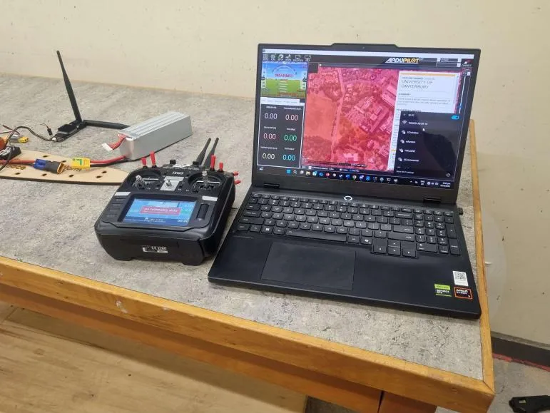 A workbench setup with a laptop running Mission Planner, a radio transmitter, a LiPo battery, an RF modem and a wiring harness laid out on a plywood board.