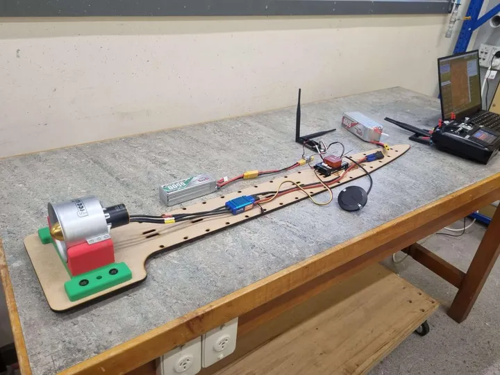 The avionics test bed laid out on a bench: a long plywood board carrying a ducted fan and motor at one end, with the ESC, battery, flight controller, RF modem and GPS puck arranged back along it.