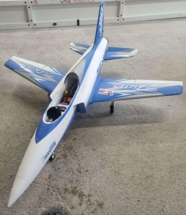 The commercial off-the-shelf electric ducted fan jet used as a dedicated avionics test platform, standing on a concrete floor with its upper hatch removed. The blue and white foam airframe has swept wings, a single swept fin and swept tailplanes, and the open bay exposes wiring and connectors ahead of the fan intake duct.