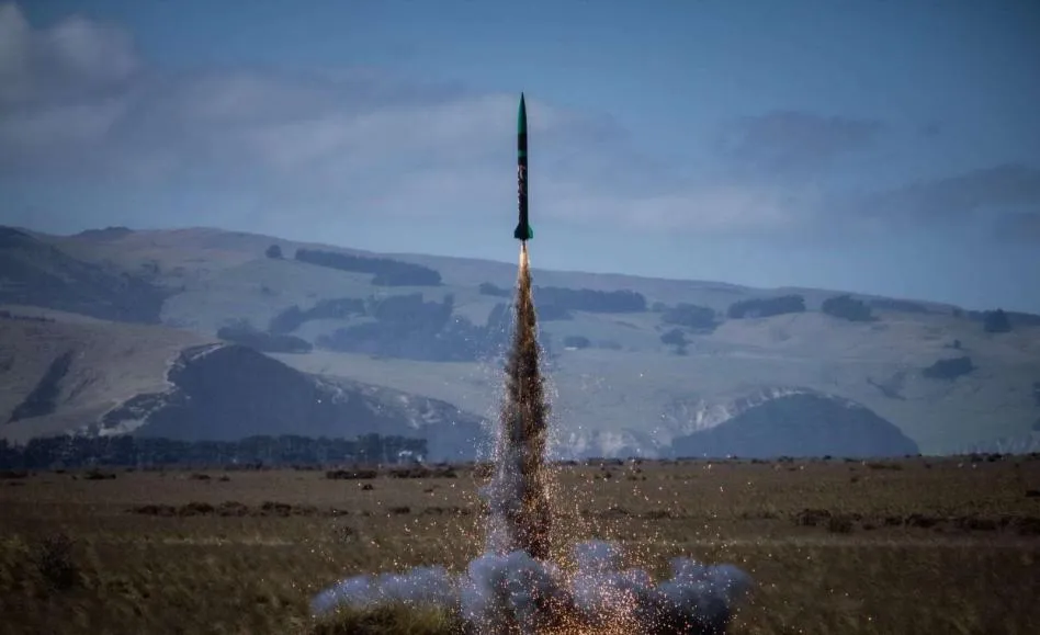 The Level 1 rocket at the moment of launch, climbing on a bright plume of sparks and smoke from the pad. Tussock farmland runs back to a line of low hills under a partly clouded sky.