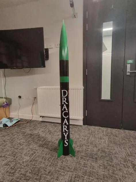 The Level 1 rocket standing on its fins indoors, roughly shoulder height. It is painted black with green bands, a green nose cone and green fins, with DRACARYS lettered down the body tube.