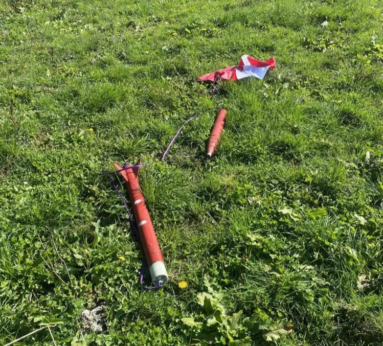 The Level 2 rocket recovered in a grass paddock after flight. The orange body tube lies intact with its fins attached, the separated nose-cone section a short distance away, and the red and white parachute laid out beyond it, still joined by shock cord.