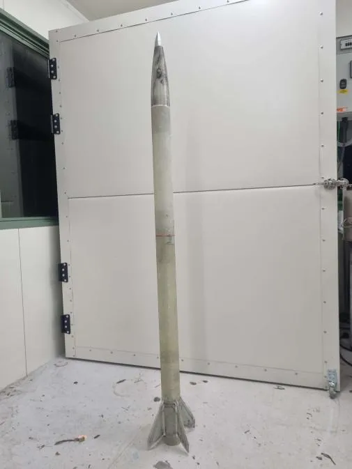 The Level 2 airframe unpainted, standing against a spray booth door. The bare fibreglass body tube runs the full height of the frame up to a long tapered metallic nose cone, with four swept fins bonded at the base.