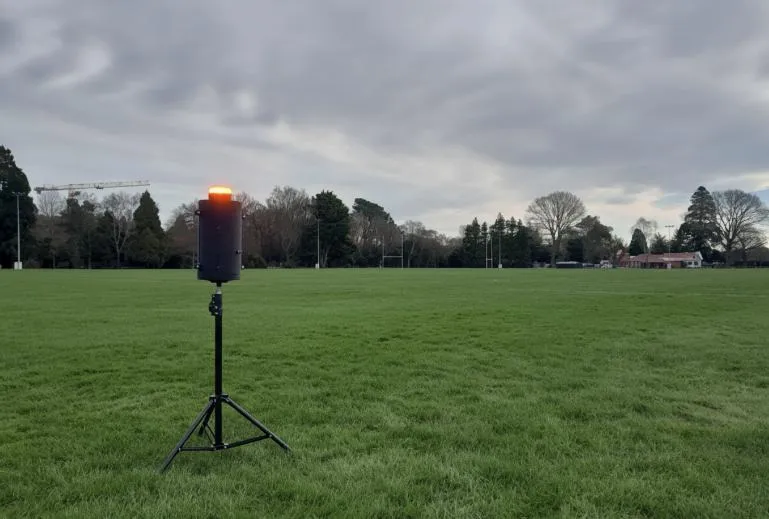 The range warning unit deployed on a sports field at dusk: a black cylindrical enclosure mounted on a tripod, its amber beacon lit at the top. Rugby posts, a treeline and a low clubhouse sit in the background under heavy overcast.