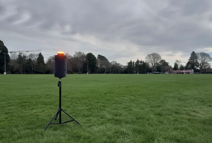 The range warning unit deployed on a sports field at dusk: a black cylindrical enclosure on a tripod with its amber beacon lit.