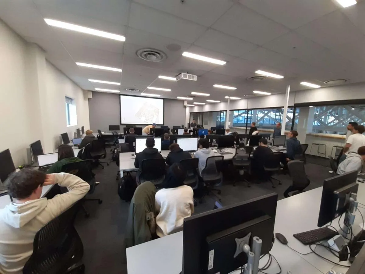 A workshop session in a university computer lab. Around twenty participants sit at rows of desktop workstations facing a projector screen at the front of the room, which shows a PCB layout. A presenter stands beside the screen.