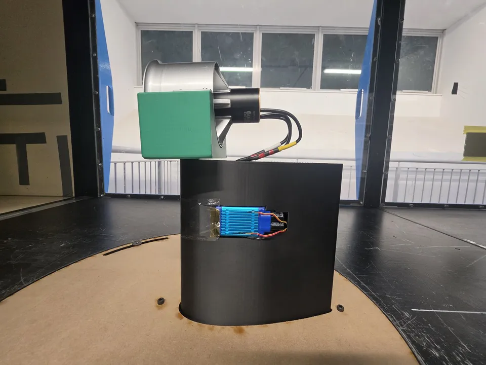 An electric ducted fan unit on the wind tunnel turntable, held above a black 3D-printed pedestal by a green printed mount. The ESC and its blue heatsink sit in a cut-out window in the front of the pedestal, with thermocouple and power leads running back from the fan to the electronics.